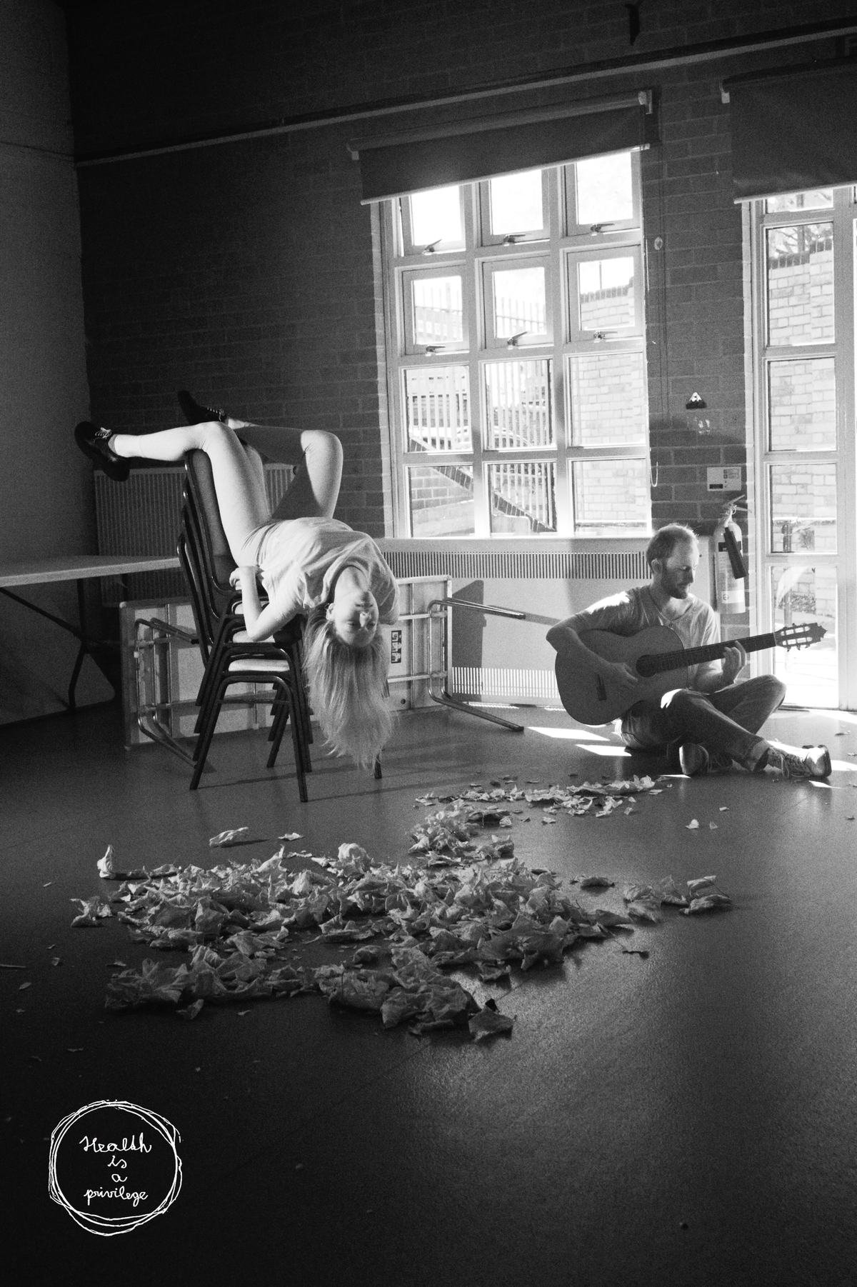 A rehearsal room strewn with dried leaves, a figure draped backwards over a chair and a second figure playing guitar by the window